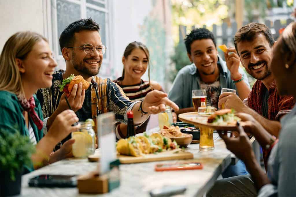 Group of friends enjoying a meal outdoors, smiling and sharing food with drinks on the table.