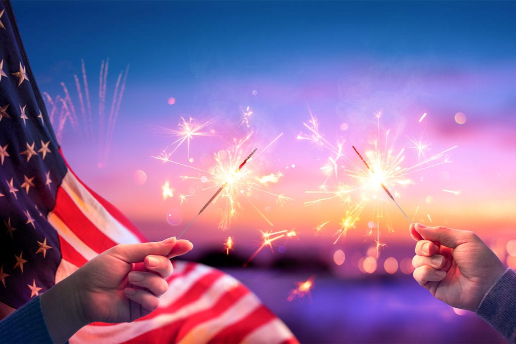 hands holding sparklers with an American flag waving behind them for the fourth of July