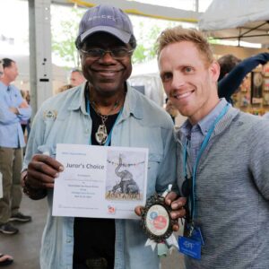 Two men pose together, one holding an award certificate and the other a medal, smiling at a lively market setting.