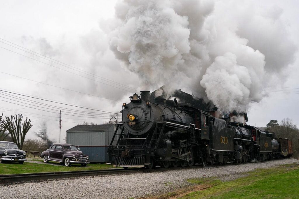 TN Valley Railroad trail blowing steam as it passes vintage cars in Chattanooga.