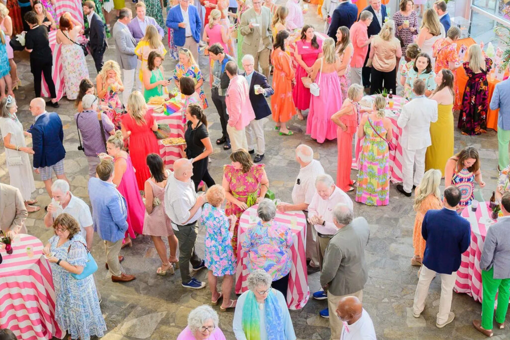 A lively outdoor gathering with people in colorful dresses and suits mingling around pink-striped tables.