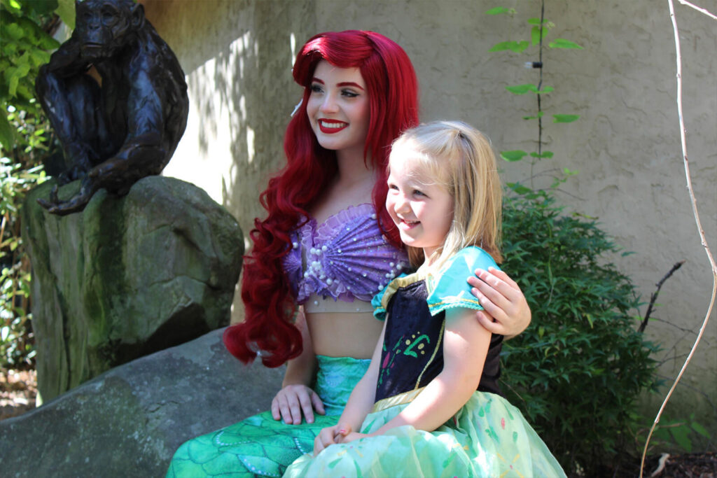 A smiling girl in a princess costume poses with a woman dressed as Ariel, surrounded by greenery and a stone statue.