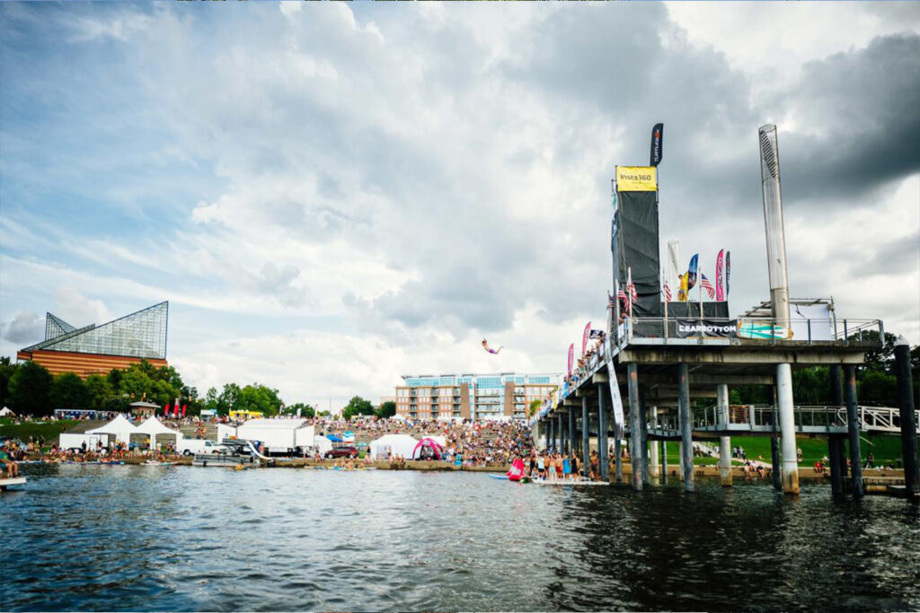 A busy waterfront scene with a pier, crowds, white tents, and a cloudy sky reflecting on the water's surface.