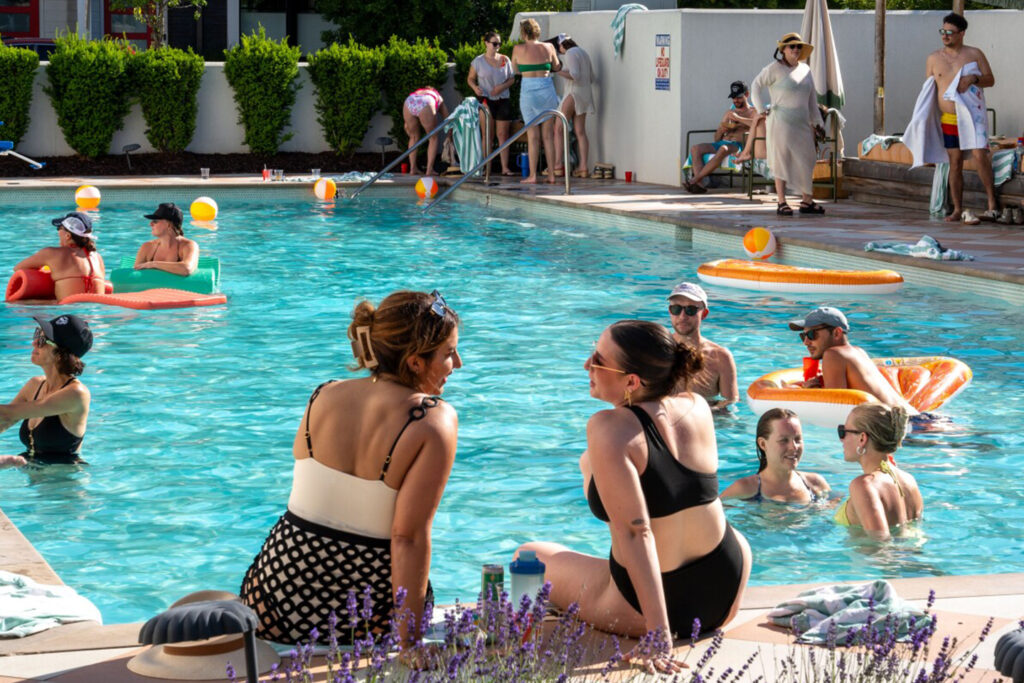 Two women in swimsuits sit near a pool, chatting while others swim and relax on inflatable floats in a sunny setting.