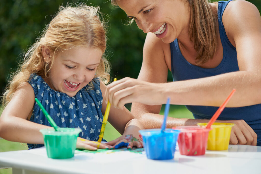 A mother and daughter joyfully painting with colorful paints on a sunny day outdoors.