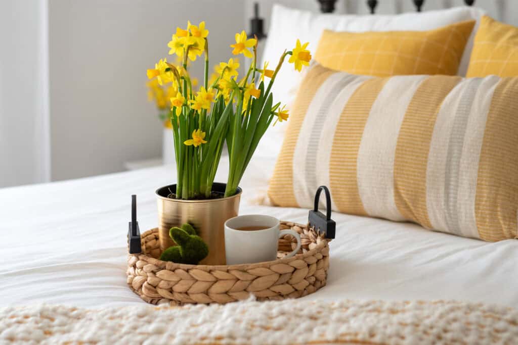 A woven tray on a bed holds yellow flowers, a small plant, and a coffee cup, with striped pillows in the background.