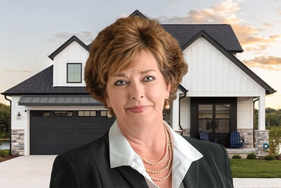 A woman in business attire stands in front of a modern house, smiling confidently against a clear sky.