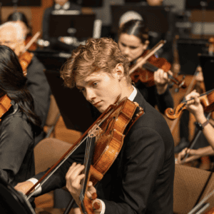 A young male musician concentrates while playing the violin in a full orchestra setting, surrounded by fellow performers.