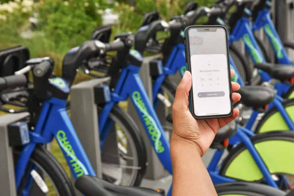 Scenic waterfront path with a blue railing, lush greenery, and fluffy clouds against a bright blue sky.
