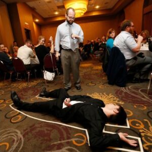 A man in a suit lies on the floor outlined in chalk while another man gestures nearby; people are seated at tables in the background.