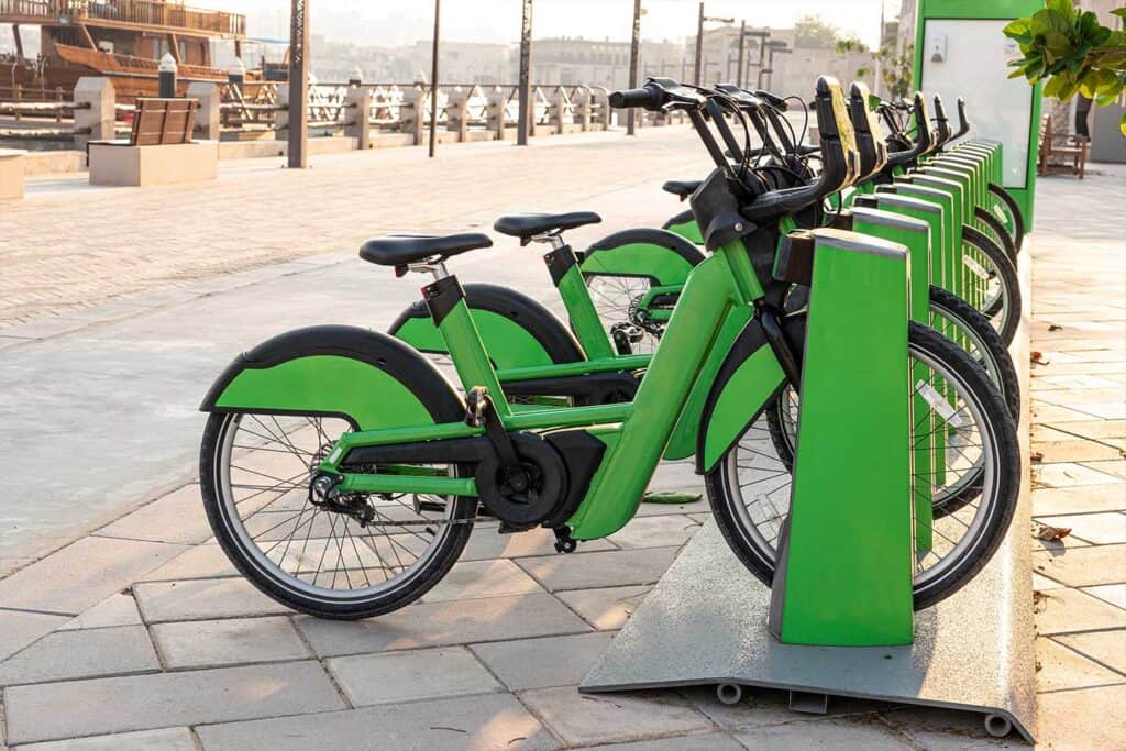 Row of green bicycles at a bike-sharing station, parked on a paved walkway with a waterfront view in the background.