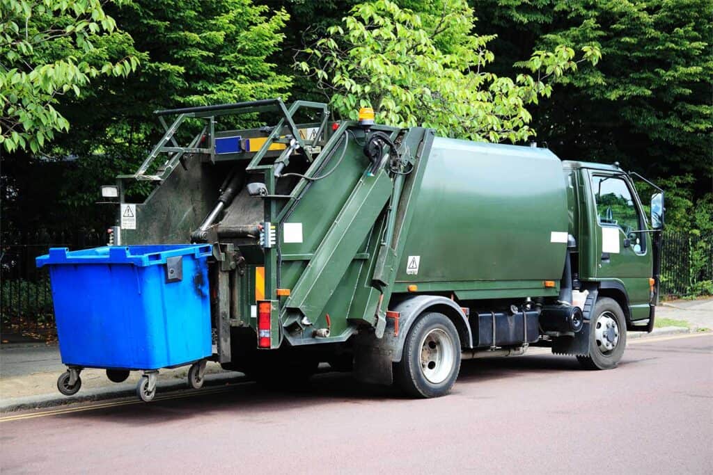 A green garbage truck parked on the street, with a blue recycling bin being emptied into the rear. Surrounding trees provide greenery.