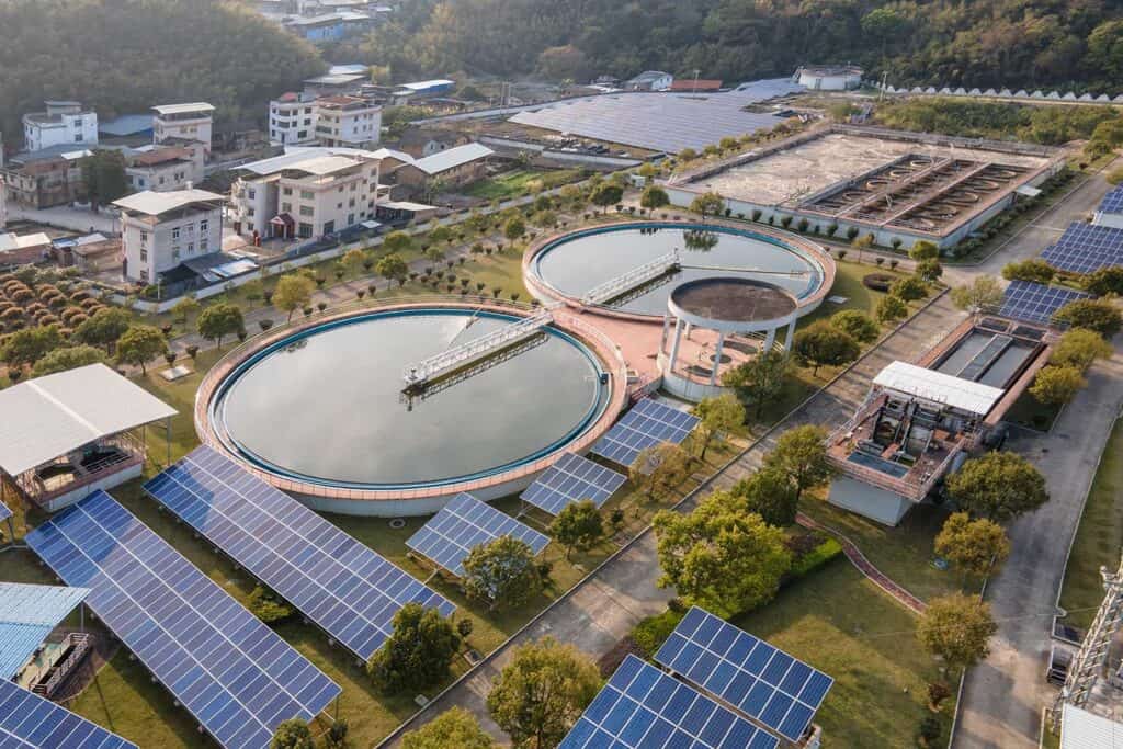 Aerial view of a solar farm with circular water treatment tanks and green spaces surrounded by buildings and hills.