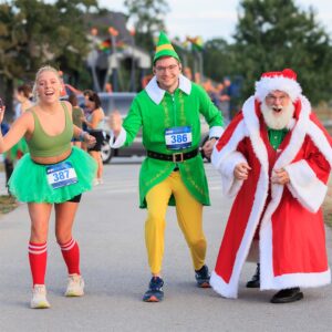A woman in a green tutu, a man in an elf costume, and Santa Claus joyfully run together in a festive race.