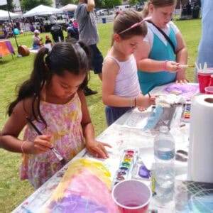 Three young girls painting colorful artwork on a sunny day at an outdoor event, surrounded by tents and grass.