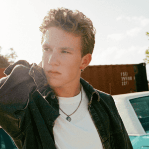 Young man with tousled hair wearing a denim jacket, standing next to an old car under soft sunlight.