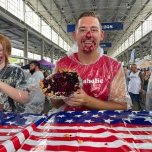Smiling man covered in pie filling holds a pie, surrounded by people at a festive event with an American flag tablecloth.
