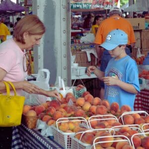 A woman and a boy examine peaches at a market stall, with colorful baskets and shoppers in the background.