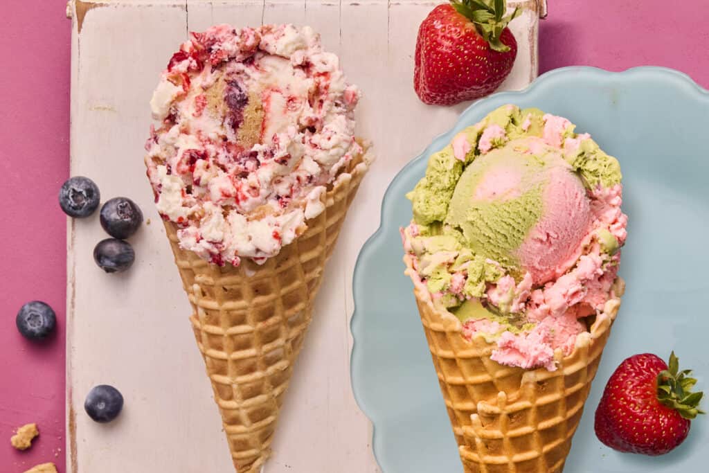 Vibrant ice cream cones with pastel scoops, one with mixed berry and the other with strawberry, set against a pink background.