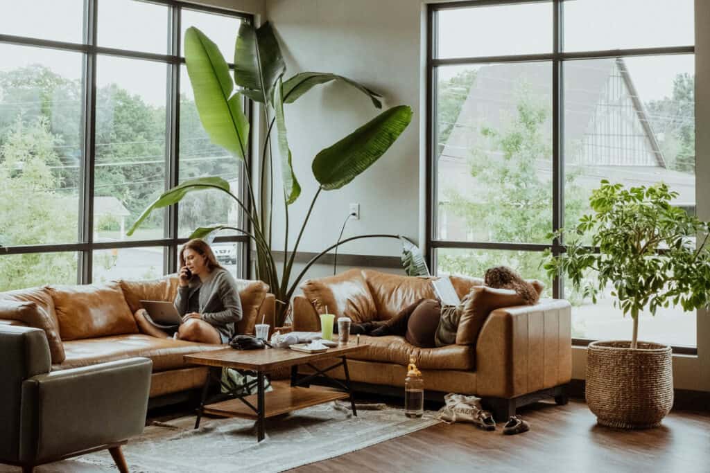 Woman sitting on a leather couch in a bright room with large windows and indoor plants, reading on a tablet.