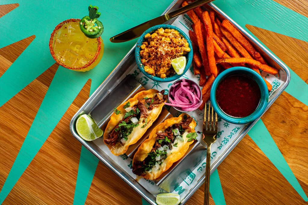 A metal tray holds two loaded tacos, sweet potato fries, corn, pickled onions, and a colorful cocktail on a vibrant table.