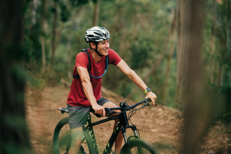 A smiling cyclist in a red shirt and helmet rides a mountain bike on a forest trail surrounded by lush greenery.