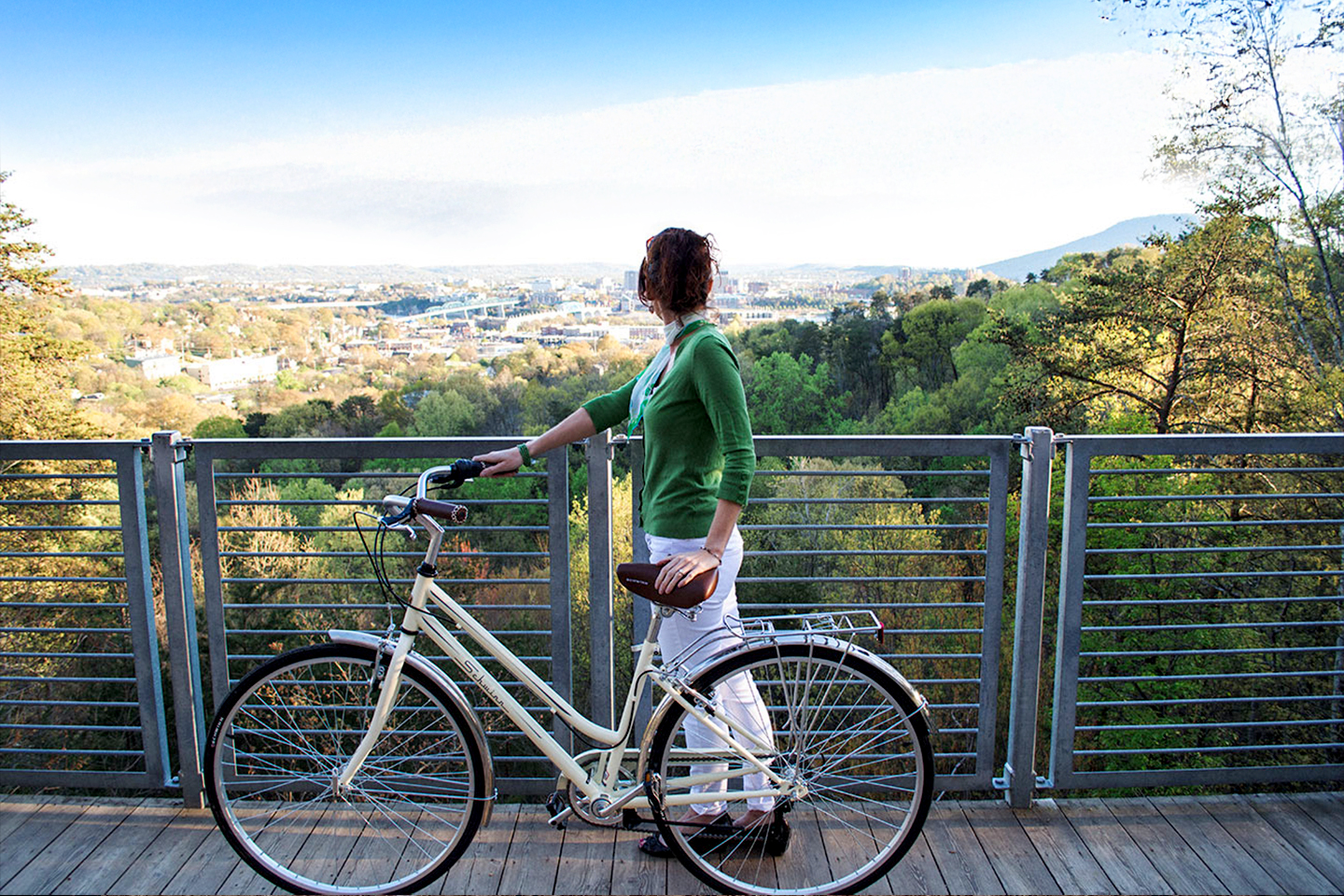 Person in a green sweater stands beside a bicycle, gazing over a scenic view of a city surrounded by greenery.