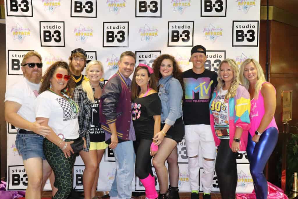 A vibrant group poses in 80s-inspired outfits against a branded backdrop, featuring bright colors and bold accessories.