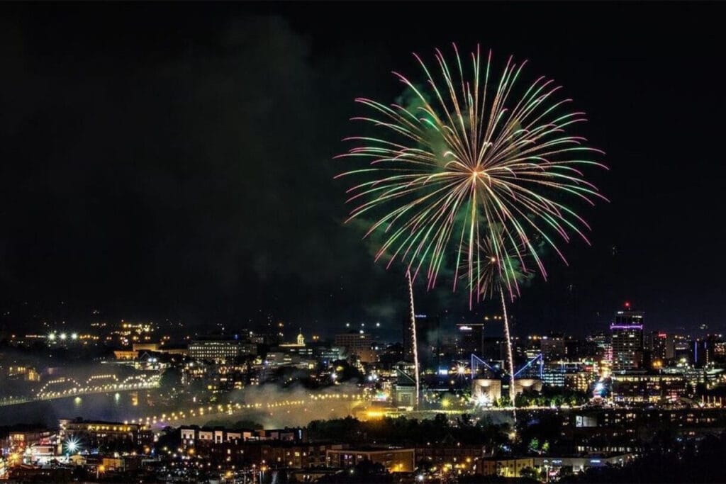 Colorful fireworks illuminate a night sky above a city skyline, with vibrant bursts reflecting over the water.
