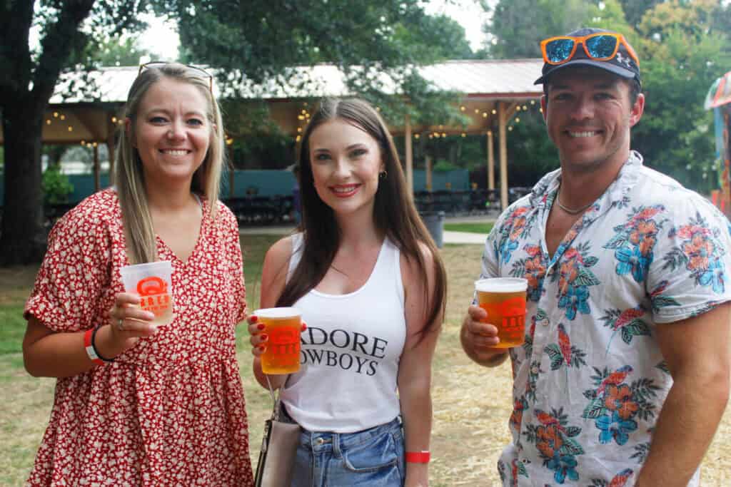 Three friends smile while holding drinks at an outdoor event, surrounded by trees and festive decorations.