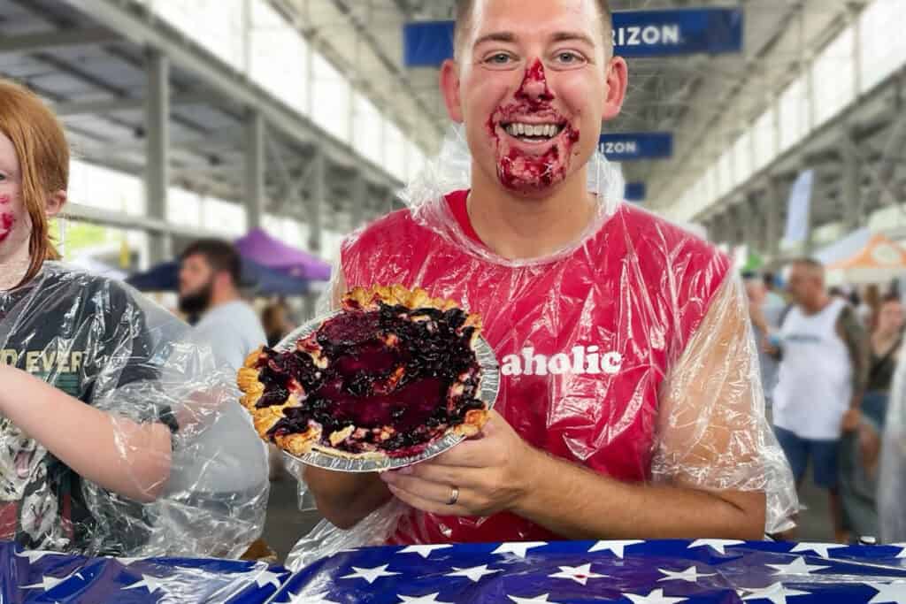 A smiling man in a red shirt holds a blueberry pie, covered in pie filling, at a lively outdoor event.