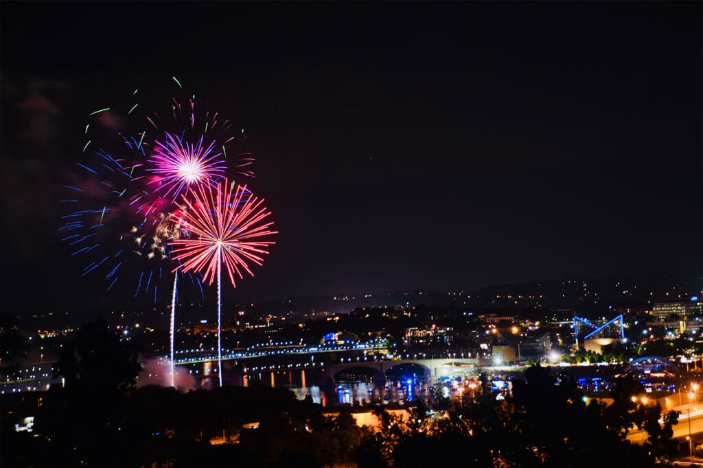 Colorful fireworks burst over a city skyline at night, illuminating buildings and a river below.