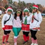 Four women dressed in festive holiday attire, including hats and green accessories, smiling together outdoors.