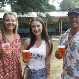 Three smiling friends enjoy drinks at an outdoor venue, surrounded by greenery and a festive atmosphere.