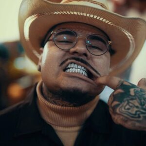 A man in a cowboy hat and glasses grins widely, revealing teeth with a mouth grill, wearing a staff badge and a turtleneck.