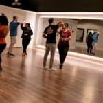 A dance studio with couples practicing various dance styles in front of a large mirror, featuring a wooden floor and soft lighting.