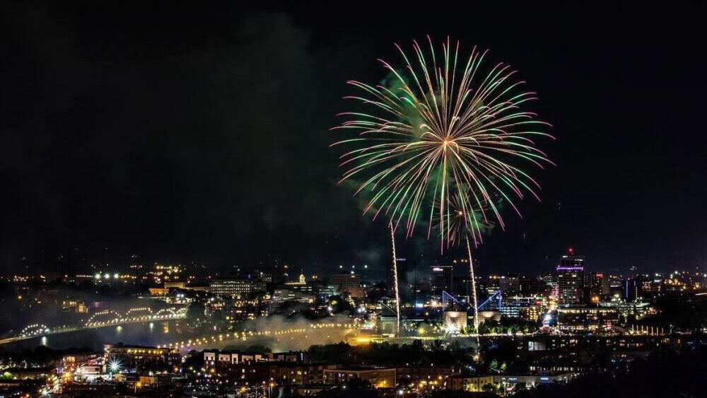 Colorful fireworks explode in the night sky above a city skyline, illuminating the buildings and reflecting off the water.