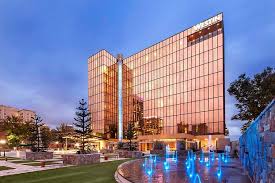 Modern glass hotel building with a reflective copper facade, surrounded by lush greenery and illuminated fountains at dusk.