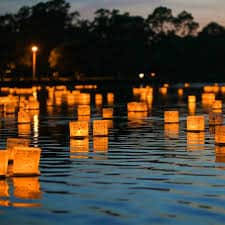 Illuminated lanterns float on a calm lake at dusk, reflecting warm light against the darkening sky and trees.