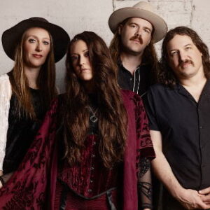 A six-member band poses against a textured wall, wearing a mix of casual and vintage clothing, with a mix of hairstyles.