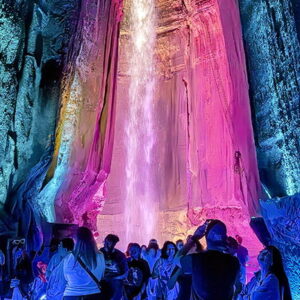 Visitors gather under a stunning, brightly lit waterfall in a dark cave, with vibrant pink and blue lights illuminating the scene.