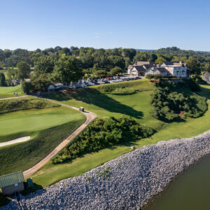 Aerial view of a golf course beside a river, featuring a green, sandy bunker, and a luxury building on a hillside.