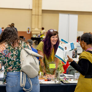 A lively event scene with a woman smiling and engaging with another attendee at a vendor booth, showcasing brochures and products.