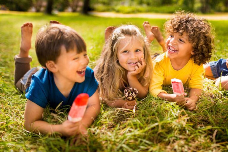 Three smiling children lie on grass, enjoying fruity popsicles, surrounded by greenery and sunlight.