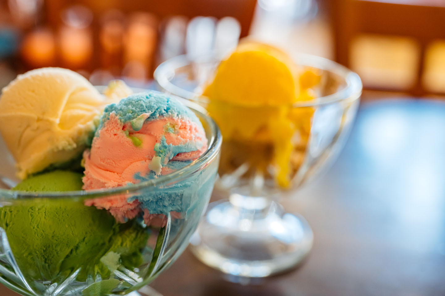 Two glass bowls filled with colorful ice cream scoops: green, pink, yellow, and white, set against a blurred background.