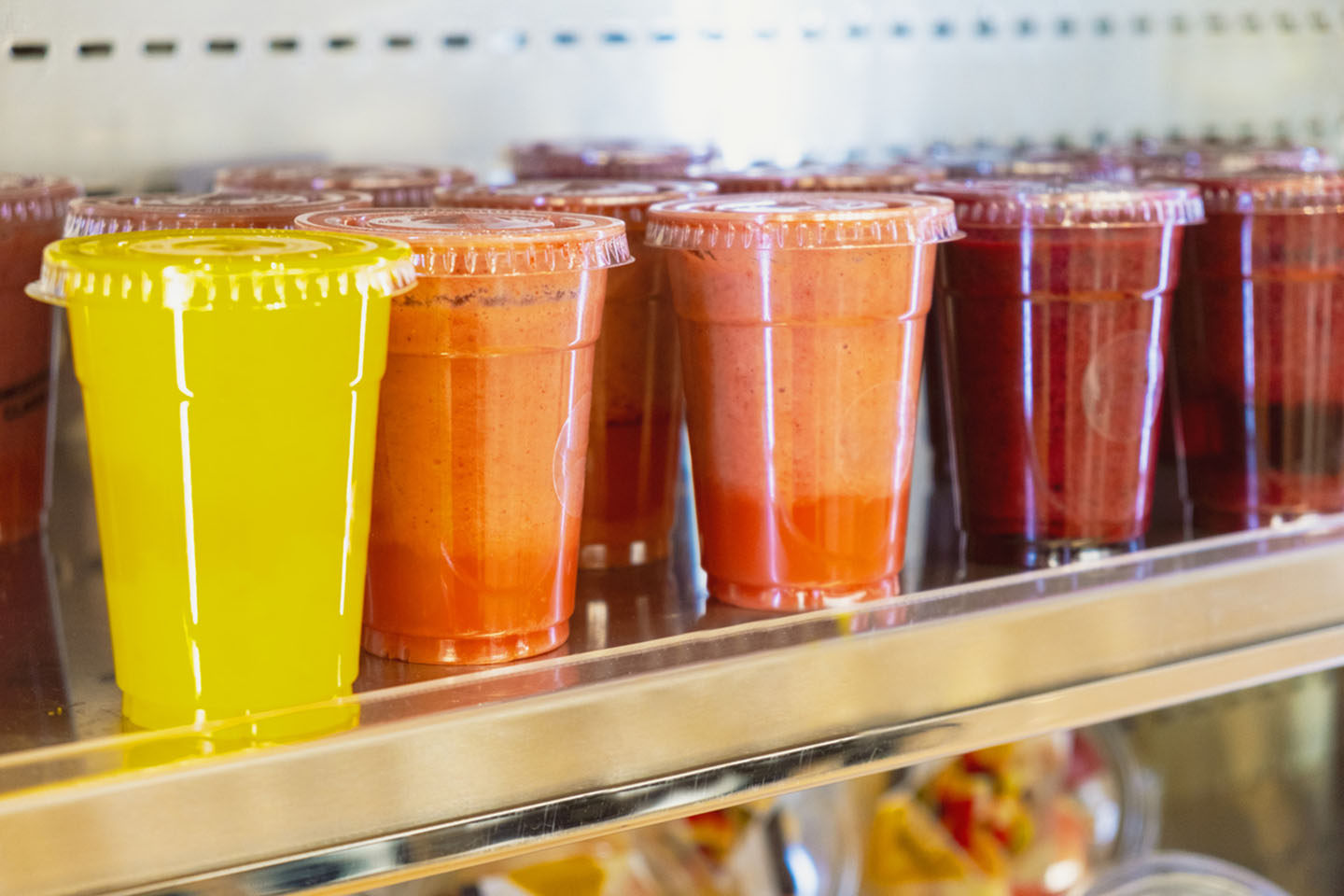 Colorful smoothie cups lined up on a shelf, featuring vibrant yellow, orange, and red beverages in clear containers.