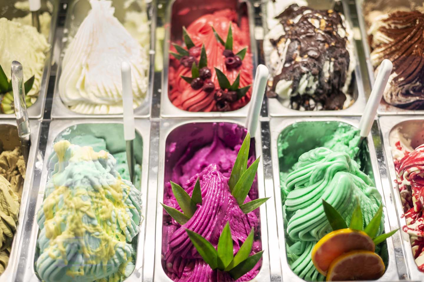 Colorful gelato varieties in metal tubs, topped with leaves and fruit slices, arranged in a tempting display.