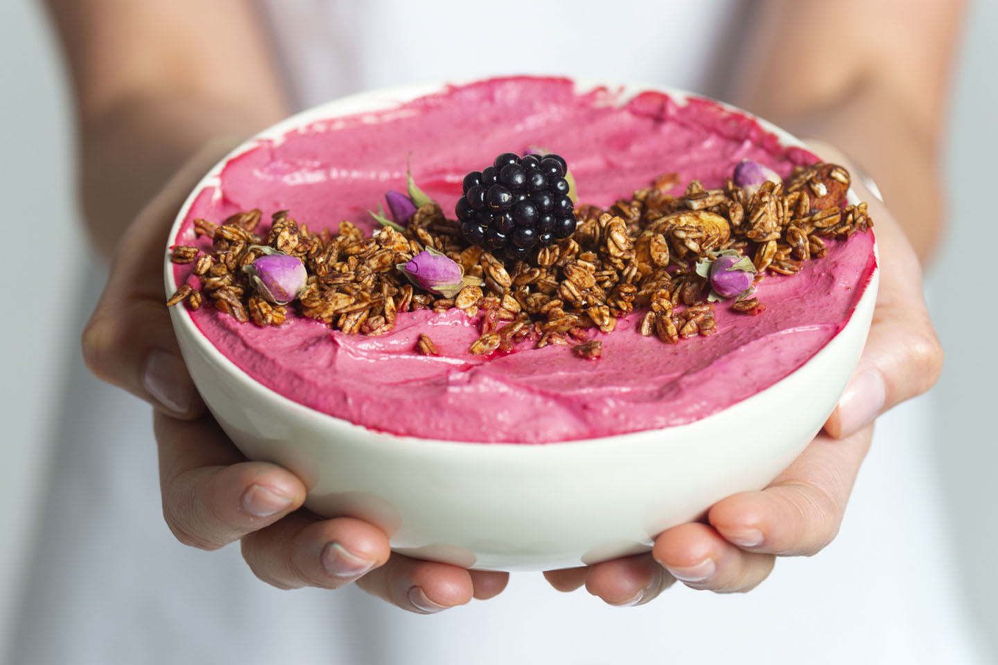A person holds a bowl of pink smoothie topped with granola, blackberries, and rose petals against a light background.