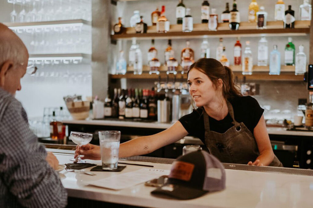 A bartender serves a cocktail to a patron at a stylish bar with various bottles displayed in the background.