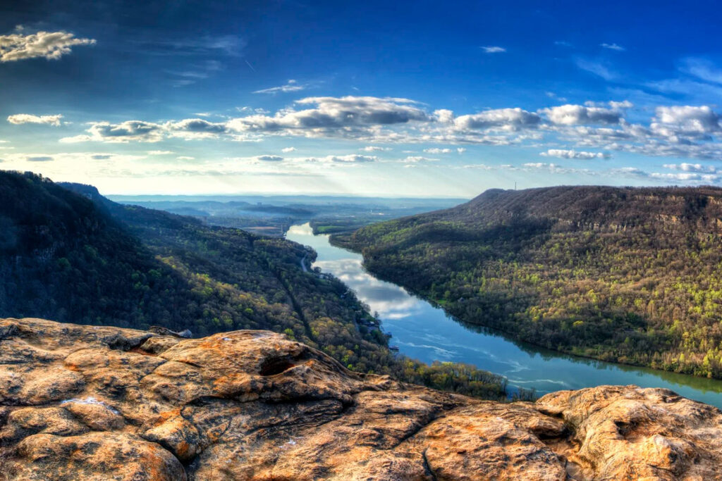 Vast landscape featuring a winding river surrounded by lush green hills under a bright blue sky with scattered clouds.
